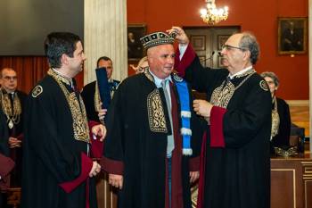 The honored SNF Co-President, Andreas Dracopoulos, standing between the Rector and the Dean of the School of Health Sciences of the NKUA. All of them are dressed in their gowns. The Dean carefully places the hat on the head of the honoree.